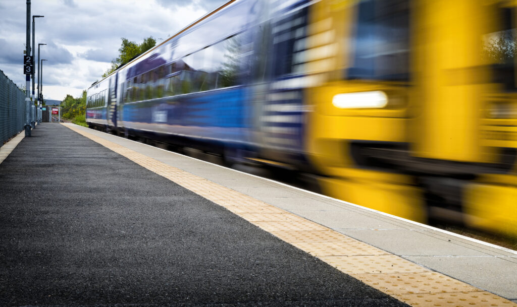 Modern train racing through railway station in Scotland with tactile flooring near the edge of the platform